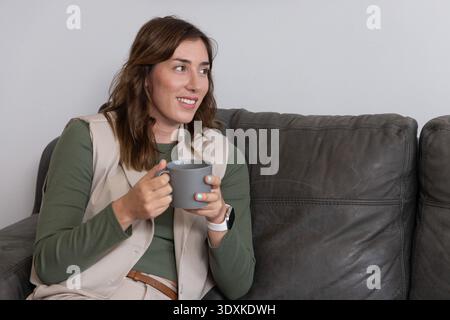 Woman sitting on gray couch holding gray ceramic mug, looking right, wearing vest and smartwatch Stock Photo