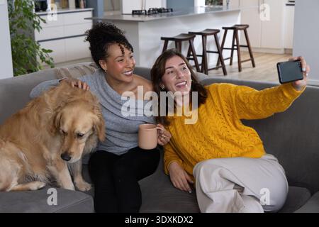 Diverse female friends on gray sectional couch at home taking phone selfie holding mug petting dog Stock Photo