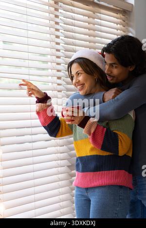 Couple embracing, pointing beside horizontal blinds, in Santa hat and sweater, holding red mug Stock Photo