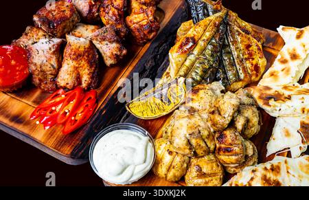 A large wooden board holds different types of grilled meat and fish along with sauce and vegetables during a meal with friends. Stock Photo