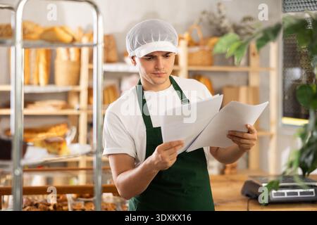 Young baker carefully reads documents and makes calculations in his head, in the interior of private bakery Stock Photo