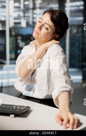 Fatigued woman in data center office relaxing muscles, stressed by long hardware troubleshooting session. Exhausted server hub worker stretching back after sitting too long at computer desk Stock Photo