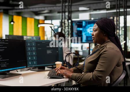 Female programmer in office using large language model application to get help with coding tasks. African American woman at work interacting with AI LLM chatbot PC GPT software on PC Stock Photo
