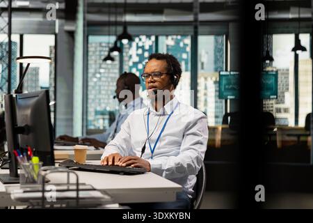 Startup helpdesk admin in office speaking in headset microphone, responding to service alerts. African american IT specialist doing tech support, helping users across remote infrastructure Stock Photo