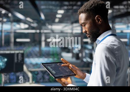 Black IT expert uses tablet to review system infrastructure on industrial platform, doing machine learning automatization. Data center technician does maintenance with artificial intelligence. Stock Photo