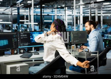 Data center female engineer stretching back to combat sedentary fatigue after working long hours at desk. African american woman in server hub fixing incorrect posture, alleviating discomfort Stock Photo