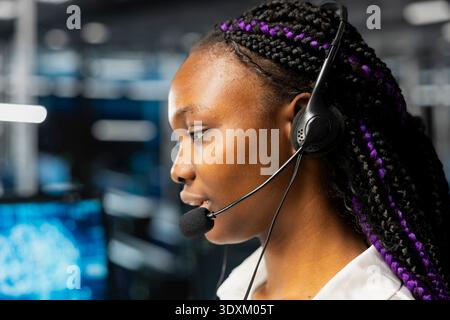 Close up of female engineer in data center speaking in headset mic, using AI to provide user technical support. Server room IT expert assisting clients during remote call using artificial intelligence Stock Photo