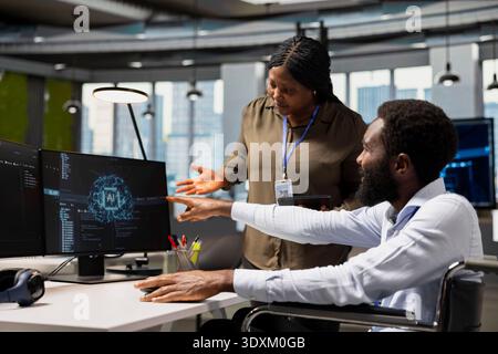 Manager overseeing tasks progress done by worker with paraplegia using AI to fix glitches. Team leader supervising programmer in wheelchair using artificial intelligence to do optimizations Stock Photo