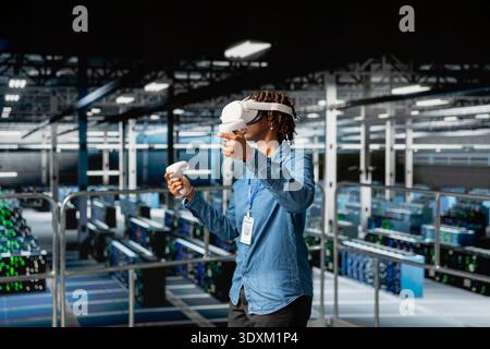 Black software engineer works with virtual reality tech on industrial platform, overseeing server racks infrastructure and AI visualization. VR equipment used in data center, computer science. Stock Photo