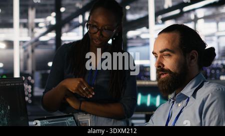 Data center system administrators working together, collaborating on neural network software development. Server hub worker and african american colleague implementing AI tech in workplace, camera A Stock Photo