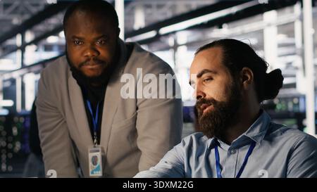 Supervisor in server room monitoring worker managing AI training datasets. African american team leader and engineer in data center supervising system operations supporting neural networks, camera A Stock Photo
