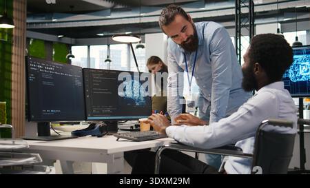 Manager supervising developer with paraplegia writing code, developing deep learning APIs. Supervisor overseeing work done by IT expert in wheelchair troubleshooting software errors using AI, camera A Stock Photo