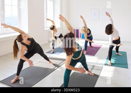 Women practicing extended side angle yoga in airy sunlit studio with mats Stock Photo