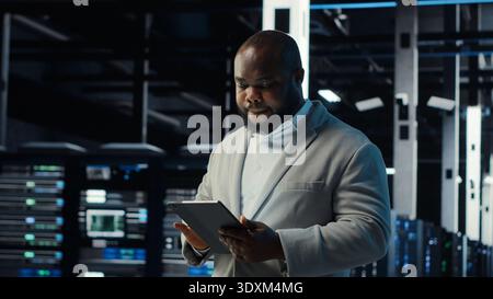 IT professional looking over data center checkup checklist paperwork on tablet. African american man walking in server farm, reading documentation files on device, camera B Stock Photo
