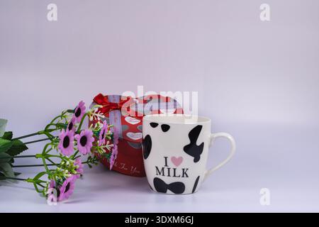 Ceramic milk mug with heart shaped gift box and purple flowers isolated on white background. Romantic celebration concept for Valentine, anniversary o Stock Photo