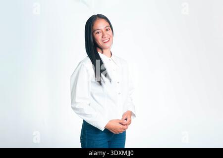 A young Indonesian woman in a white button-up shirt posing with crossed arms and a confident smile, isolated on a white studio background. Stock Photo