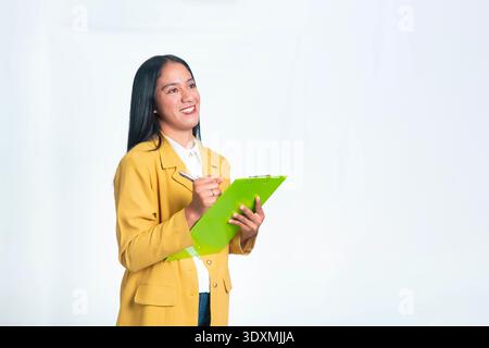 A happy Indonesian woman in a yellow blazer smiling while writing notes on a green clipboard, isolated on a white background. Stock Photo