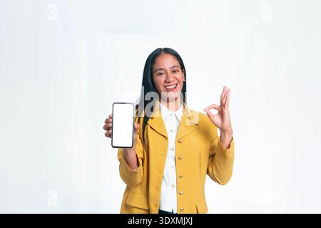 A joyful Indonesian woman in a yellow blazer holding a smartphone with a blank white mockup screen while giving an OK hand sign on a white background. Stock Photo