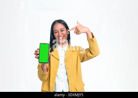 A smiling Indonesian woman in a yellow blazer holding a smartphone with a green screen chroma key mockup while pointing at it on a white background. Stock Photo