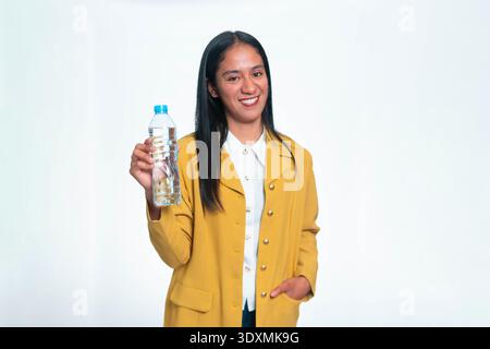 Portrait of a happy Indonesian woman wearing a yellow jacket and holding a plastic mineral water bottle, isolated on white background. Stock Photo