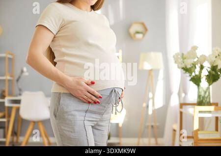 Pregnant woman standing in cozy home, holding belly with both hands and enjoying maternity moments. Stock Photo