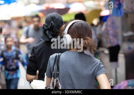 Woman with brown hair in bun wearing headphones shopping in outdoor market, dynamic street life, rear view, at Wang Lang Market. Stock Photo