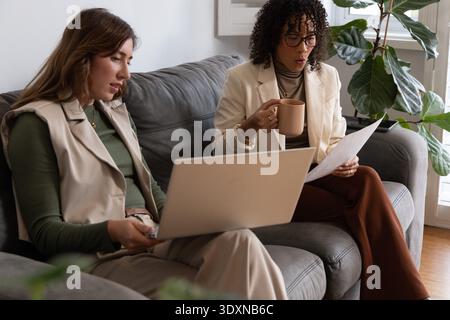 Diverse female coworkers in blazers reviewing documents on laptop at home on gray sofa, tan mug Stock Photo