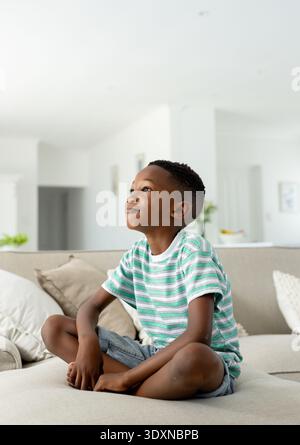 African American boy sitting on light sofa at home, in striped-tee, gazing near pillows fruit-bowl Stock Photo