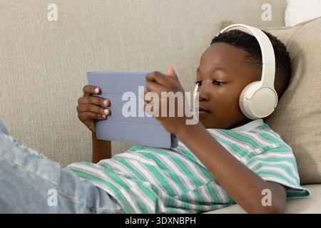 African American child reclining on sofa holding grey tablet wearing green shirt and cream headset Stock Photo
