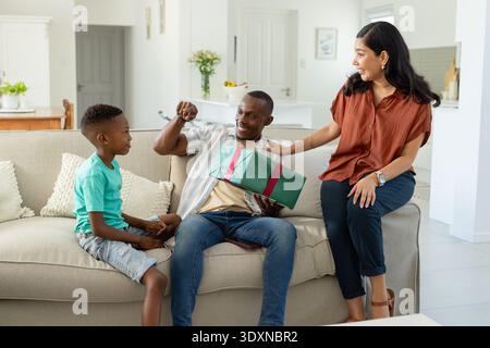 Diverse family sitting on beige sofa, father holding green wrapped gift with red ribbon, smiling Stock Photo