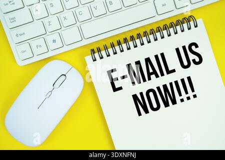 Notepad with the text "E-Mail Us Now!!!" with white keyboard and wireless mouse on yellow background Stock Photo
