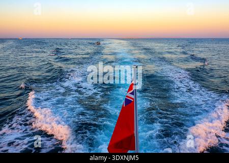 Golden sunset paints vast ocean waves. Red flag flaps in the salty breeze. White wake trails behind moving vessel. Distant ships dot the calm horizon. Stock Photo