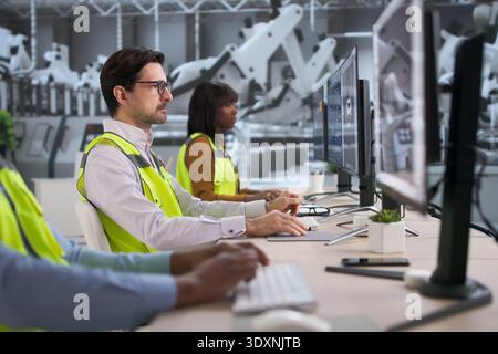 Diverse Team Of Engineers And Technicians Using Computers Near Car Assembly Line. Young Software Developers Stock Photo
