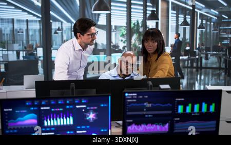 Diverse Team Of Financial Analysts Uses Computers And Data Screens, Predicting Market Trends And KPIs, Collaborating Stock Photo