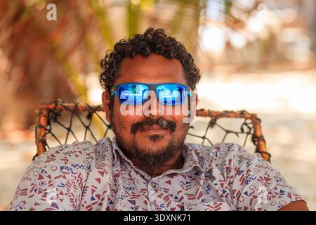 Man relaxes on beach chair with blue sunglasses. Curly hair and beard frame his calm, content expression. Patterned shirt adds color to tropical backd Stock Photo