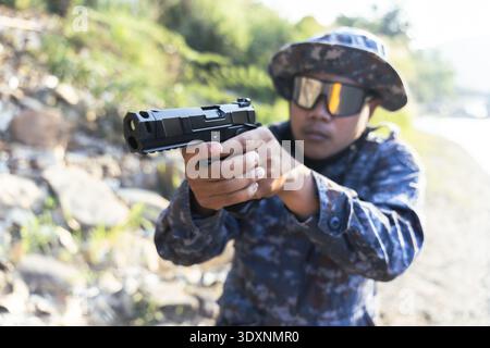 Focused Asian man in camouflage uniform and hat aiming handgun. serious soldier with tactical sunglasses practices shooting at an outdoor range with d Stock Photo