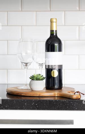 Red wine bottle mockup with two empty glasses and a succulent on a wooden board in a modern kitchen Stock Photo