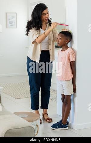 Diverse adult woman measuring youth boy's height against white wall using pink pencil and level Stock Photo