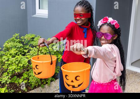 Two African American female children standing on porch holding treat buckets, wearing red mask Stock Photo
