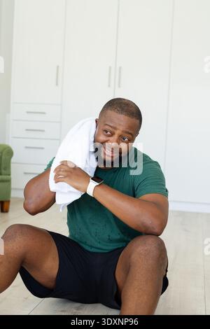 Man sitting on floor wiping sweat with white towel at home, wearing green tee and smartwatch Stock Photo