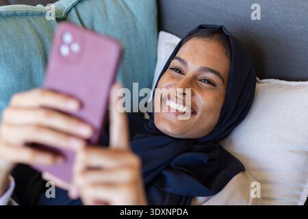 Woman reclining on sofa holding mauve phone, wearing dark hijab, shades, teal cushion, light pillow Stock Photo