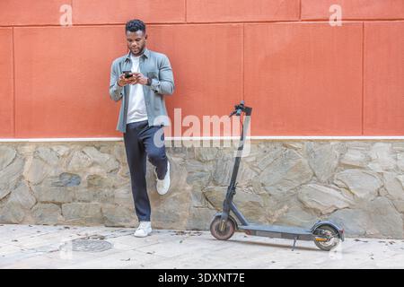 A young man in casual attire relaxes against a vibrant orange wall, checking his smartphone. Next to him, an electric scooter stands ready for use, re Stock Photo