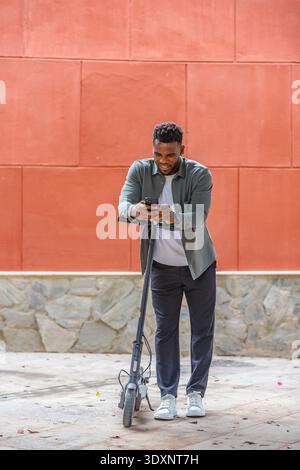 A young man stands beside his scooter, focused on his smartphone while smiling. The vibrant red wall and stone pavement create a lively urban atmosphe Stock Photo