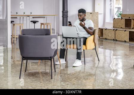 A young man sits comfortably in an elegant chair, grimacing angrily and concentrating on his laptop while occasionally consulting his phone in a conte Stock Photo