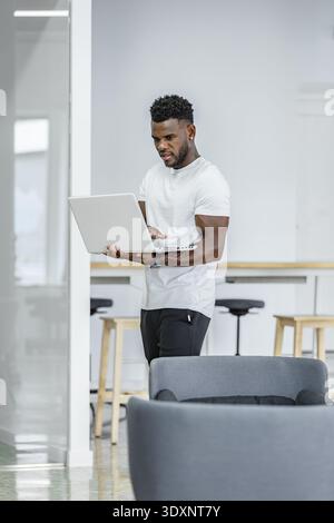 A young man stands while focused on his laptop in a contemporary office. The bright environment features stylish furniture, promoting a productive atm Stock Photo