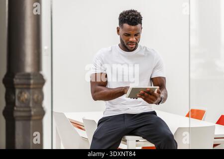 A 30-year-old African-American man is seated at a table in a bright, contemporary coworking office. He is focused on using a tablet, showcasing a blen Stock Photo