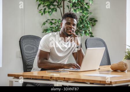 A young man is focused on his laptop, sitting at a wooden desk in a contemporary office filled with greenery. He appears relaxed and engaged in his wo Stock Photo