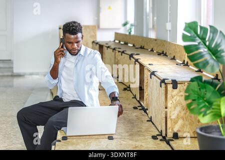 A young man is focused on a business call while sitting at a wooden table and using his laptop in a contemporary office space. Green plants add a touc Stock Photo