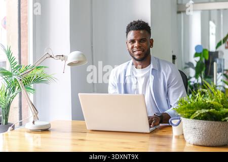 A young man sits at a wooden desk, focused on his laptop, in a bright office filled with greenery. Sunlight streams through the large windows, creatin Stock Photo