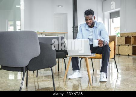 A young man in casual attire works intently on his laptop at a stylish office lounge, sipping coffee. The bright space features modern furniture and a Stock Photo